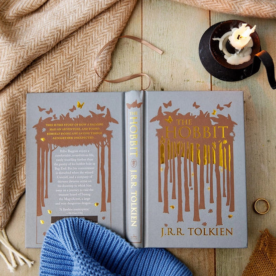 A grey hardcover book, opened and face down on a wooden table. The front cover depicts a minimalist drawing of a forest in brown. Gold text says "The hobbit." Next to the book is a blanket, a candle in an iron holder, and a gold ring.