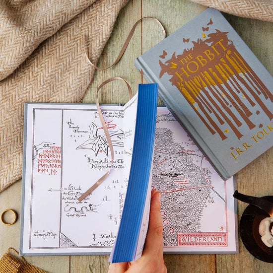 A grey hardcover book, opened and face up on a wooden table. The front cover depicts a minimalist drawing of a forest in brown. Gold text says "The hobbit." The inside cover features a drawing of a map depicting Middle Earth. Next to the book is a blanket, a candle in an iron holder, and a gold ring.