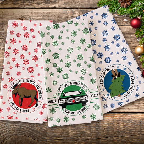 A set of three tea towels each with a different design: a moose encircled with "not a creature was stirring not even a moose," an impala car with antlers and a red button nose encircled with "Deck the halls with salt and iron impala lalala", and a Castiel sitting on top of a Christmas tree encircled with "I belive in the little tree topper". The towels have little snowflakes and anti-possession symbols adorning them. Under the towels is a wood floor, with a christmas tree at the edge.