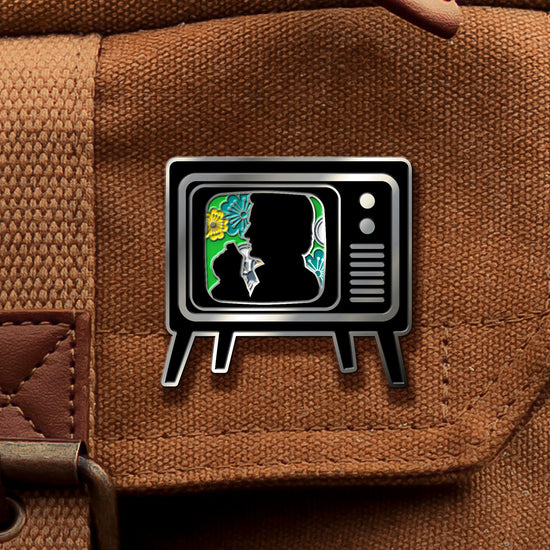A brass pin in the shape of an old TV set, on a canvas tan bag. On the screen is a black silhouette of the Trickster character from Supernatural, surrounded by green and yellow flowers.