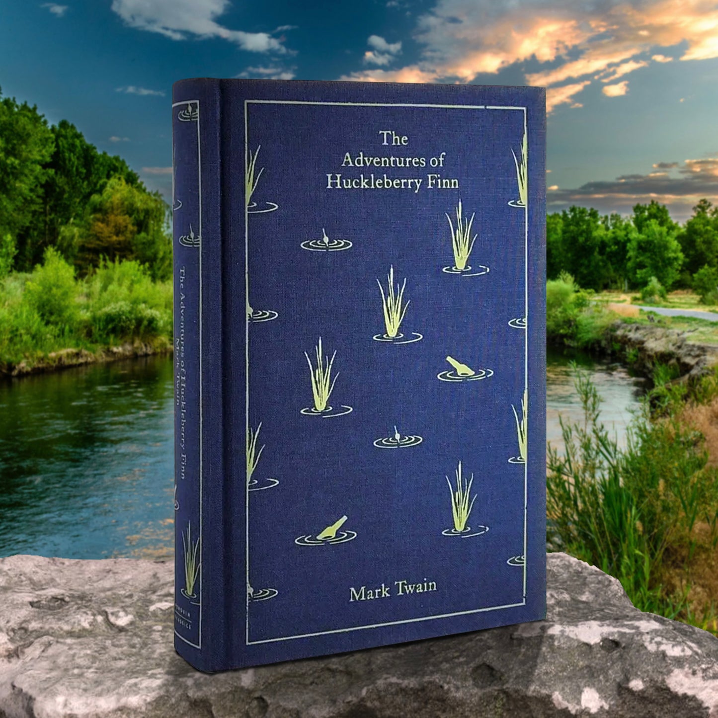 A blue book standing on a rock, in front of a river. On the cover are yellow drawings of river grass and glass bottles floating in the water. Yellow text says "The Adventures of Huckleberry Fin, Mark Twain."