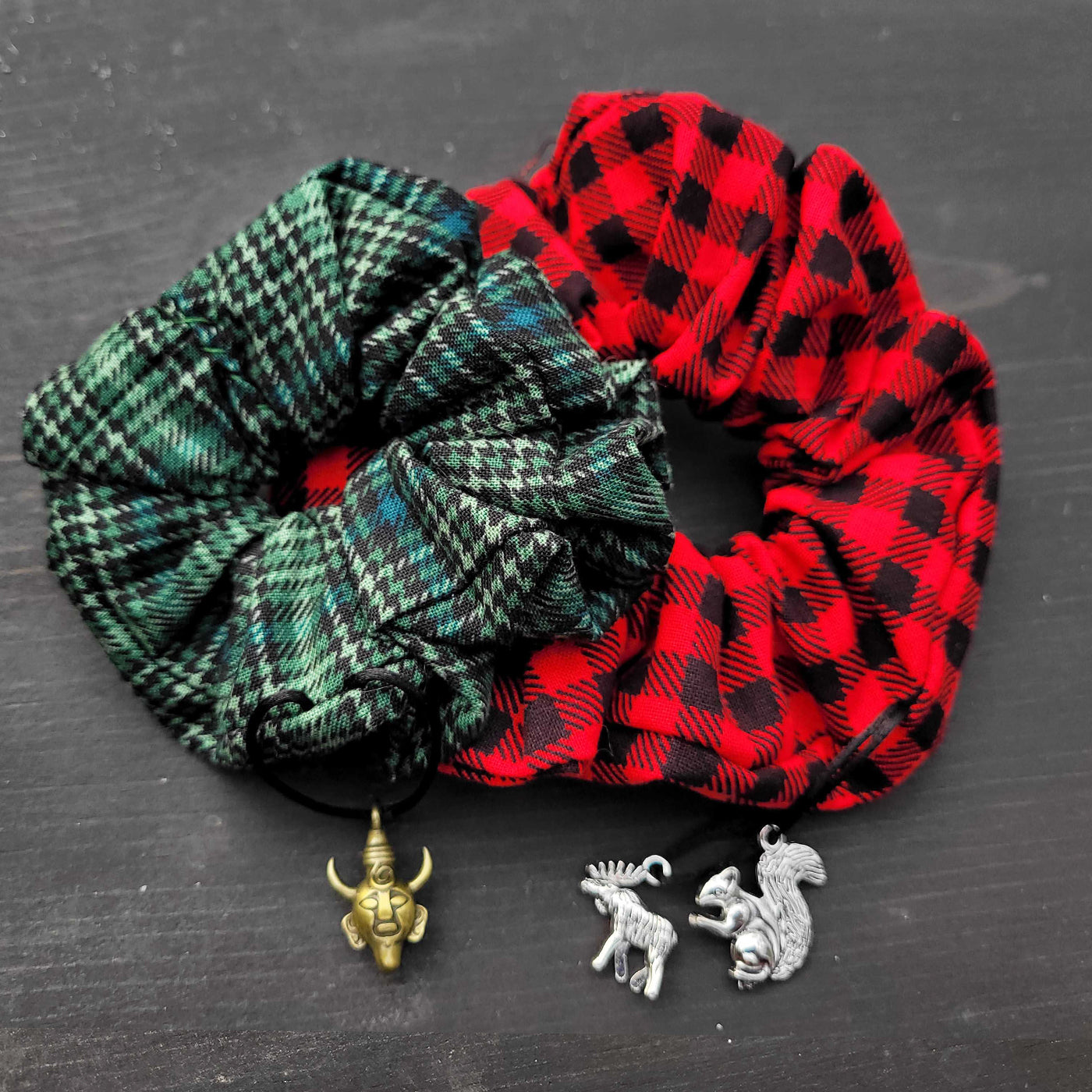 Close up view of two scrunchies against a black table top. One scrunchie has a green plaid pattern, with a brass Samulet charm at the bottom. The other scrunchie has a black and red plaid pattern, with moose and squirrel charms at the bottom.