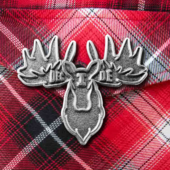Close up view of a silver pin against a red plaid background. The pin is shaped like a moose head, with "I Decide" stamped across the center of the antlers.