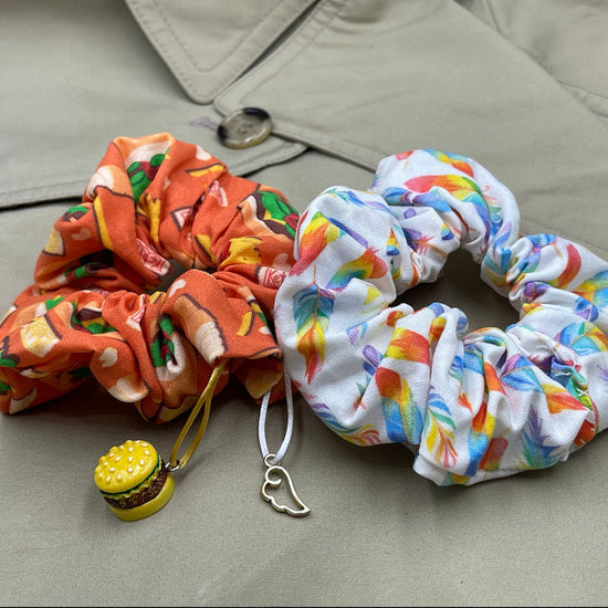 Two scrunchies side by side against a tan raincoat. The left scrunchie is orange with cartoon drawings of cheeseburgers in a pattern. A cheeseburger charm is attached to the fabric. The right scrunchie is white with rainbow colored feathers in a pattern. A silver angel wing charm is attached to the fabric.