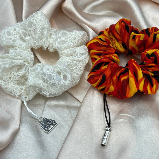 Two scrunchies side by side against white silk robe. The left scrunchie is white with a lace pattern. A silver charm shaped like open flames is atatched to the fabric. The right scrunchie is red with a flame pattern. A silver charm shaped like a fire extinguisher is attached to the fabric.