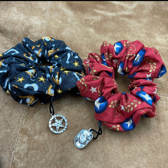 Two scrunchies side by side against a brown leather jacket. The left scrunchie is blue with horseshoes in a pattern. A charm shaped like a sheriff's badge is attached to the fabric. The right scrunchie is red with a pattern of sheriff badges and laurels. Attached to the fabric is a charm shaped like a cowboy hat.