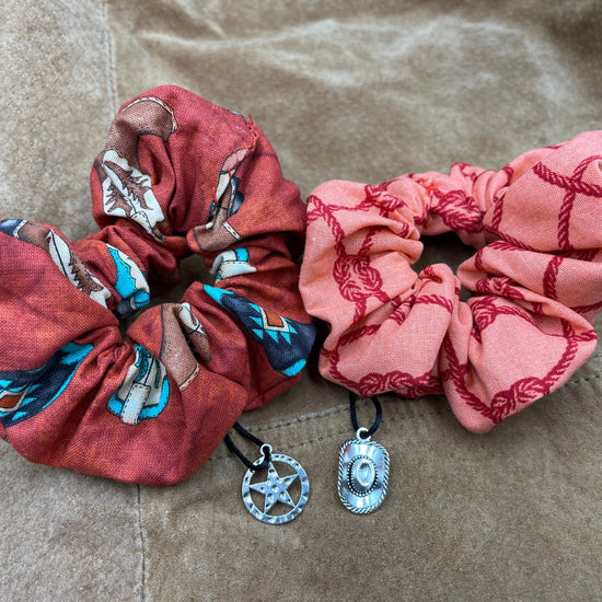 Two scrunchies side by side against a brown leather jacket. The left scrunchie is reddish brown with a pattern a cowboy boots. A charm shaped like a sheriff's hat is attached to the fabric. The right scrunchie is red with a pattern of knotted ropes. Attached to the fabric is a charm shaped like a sheriff's badge.