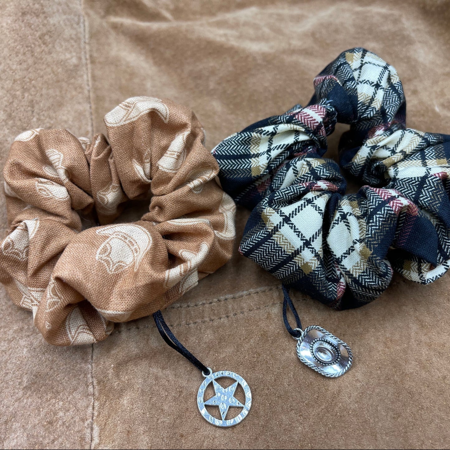 Two scrunchies side by side against a brown leather jacket. The left scrunchie is light brown with a pattern a cowboy hats. A charm shaped like a sheriff's badge is attached to the fabric. The right scrunchie is a black, white, yellow, and red plaid. Attached to the fabric is a charm shaped like a sheriff's hat.