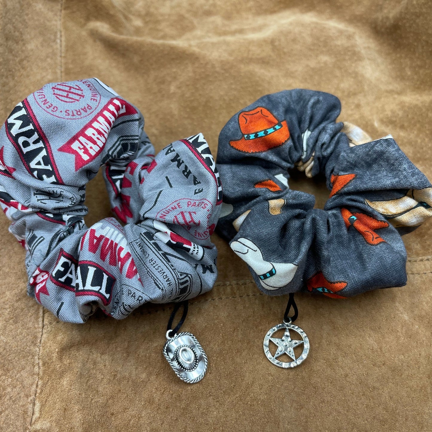 Two scrunchies side by side against a brown leather jacket. The left scrunchie is grey with a pattern a farmail logos. A charm shaped like a cowboy hat is attached to the fabric. The right scrunchie is black with a pattern of cowboy hats. Attached to the fabric is a charm shaped like a sheriff's hat.