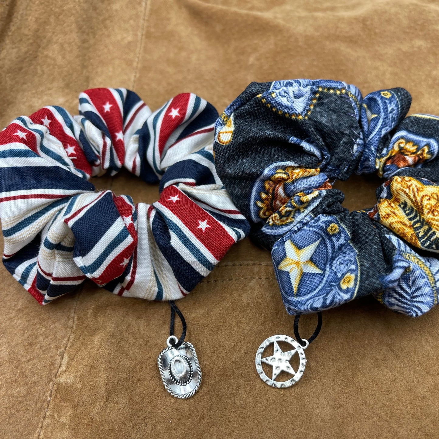 Two scrunchies side by side against a brown leather jacket. The left scrunchie has a red white and blue pattern with stars. A charm shaped like a cowboy hat is attached to the fabric. The right scrunchie is blue with a pattern of sheriff badges. Attached to the fabric is a charm shaped like a sheriff's badge.