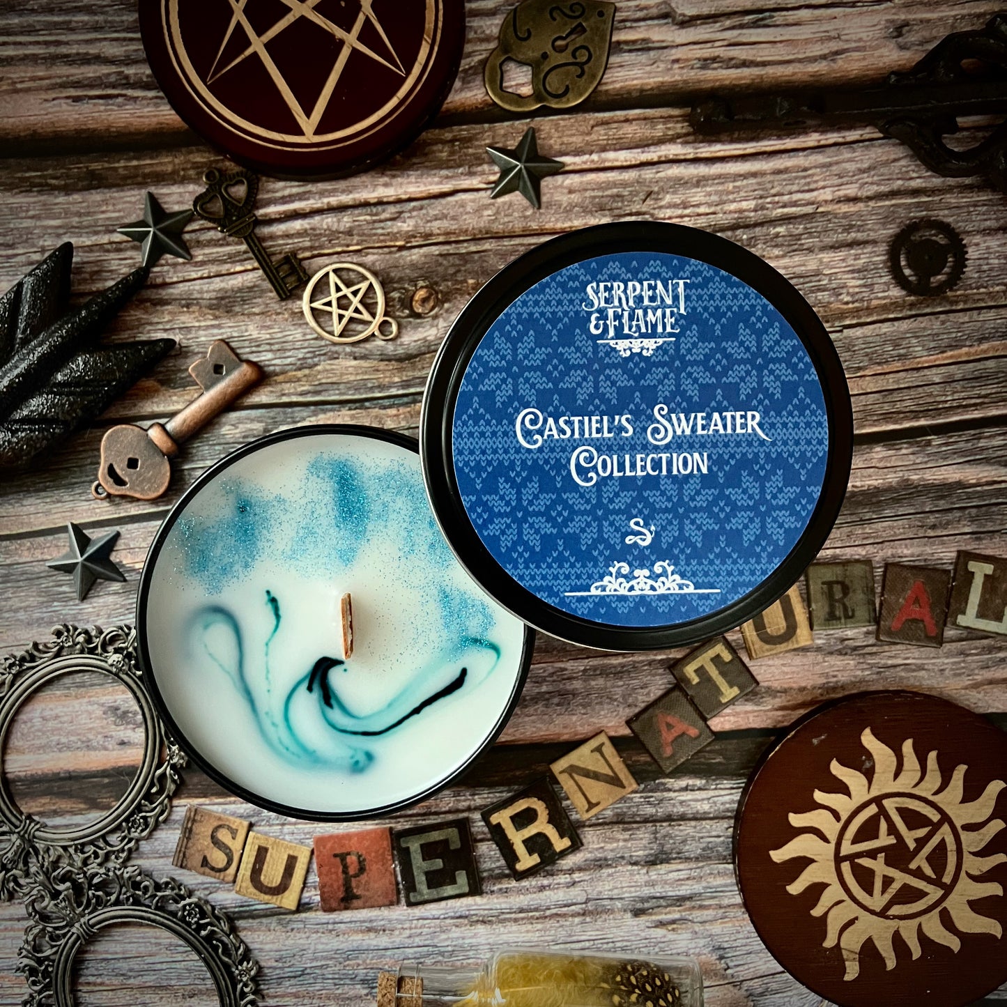 Load image into Gallery viewer, A round white candle in a blue container, sitting on a wooden table top. The lid is sitting next to the candle. The label has a light blue floral pattern, with white text saying &amp;quot;Castiel&amp;#39;s Sweater Collection.&amp;quot; Surrounding the candle are various bracelets, keys, and magical items. 
