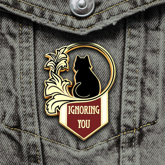 Close up view of a black, gold, amd maroon pin on a white background. The pin depicts the back of a black cat, sitting inside a circle made of flower petals. Under the cat is a maroon triangular banner with gold text that says "Ignoring you." The pin is pictured on a dark denim jacket front pocket. 