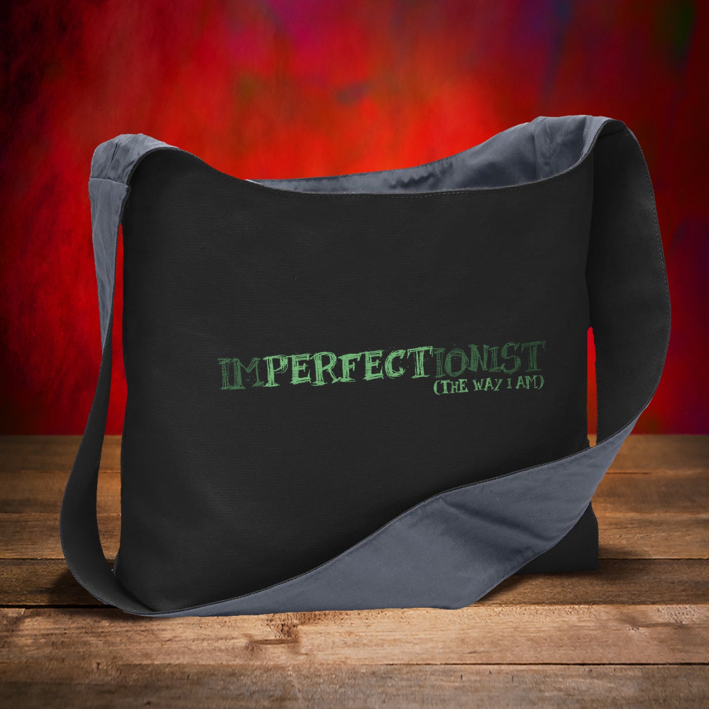 A black canvas sling bag on a wooden table, against a red background. On the front is green text saying "imperfectionist (the way I am)"