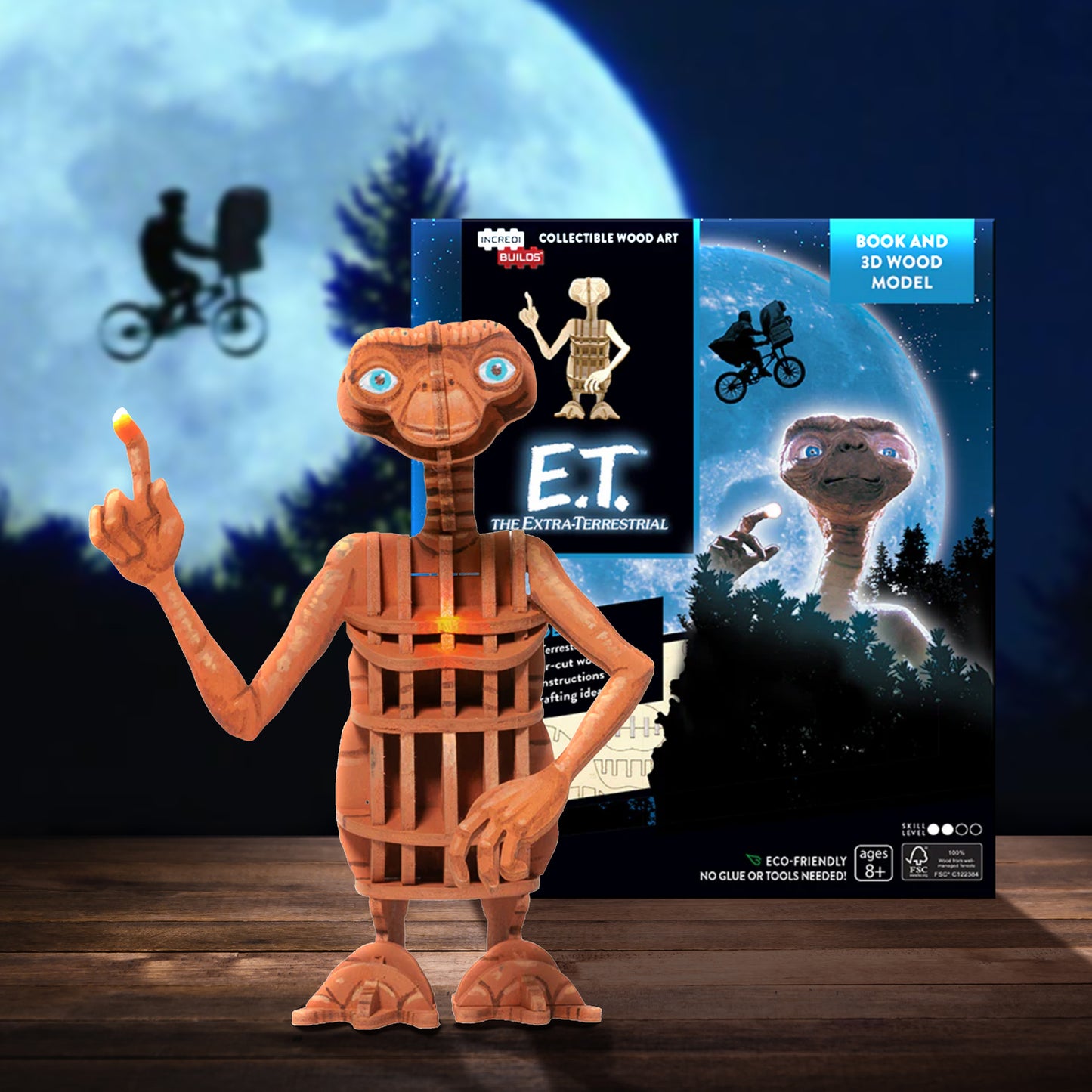 Wooden E.T. figure with packaging in the background.Behind them is the moon with a bicycle flying across the front.