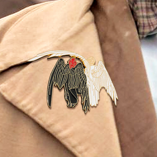 Close up view of a black and white enamel pin, attached to a light tan lapel. The pin depicts two angels side by side. The left angel is black with red hair. The right angel is white with blonde hair. The white angel's wing is slung over the black angel's shoulder