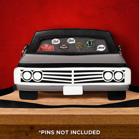 A black handbag shaped like the front of a 1967 Chevy Impala, on a wood table. Various pins line the front windshield. In the background is a red wall. At the bottom of the image is white text saying "pins not included."