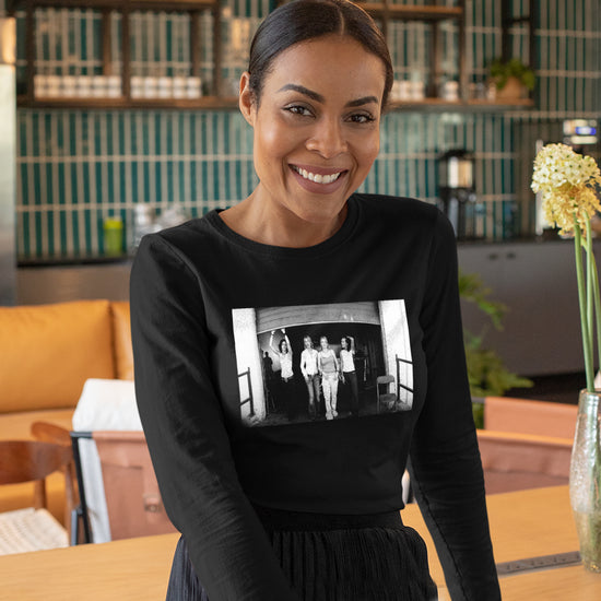 A female model wearing a black longsleeve T-shirt. On the fron of the shirt is a black and white image of four women standing in front of an open garage door. Behind the model is a lounge area with couches and a coffee station.