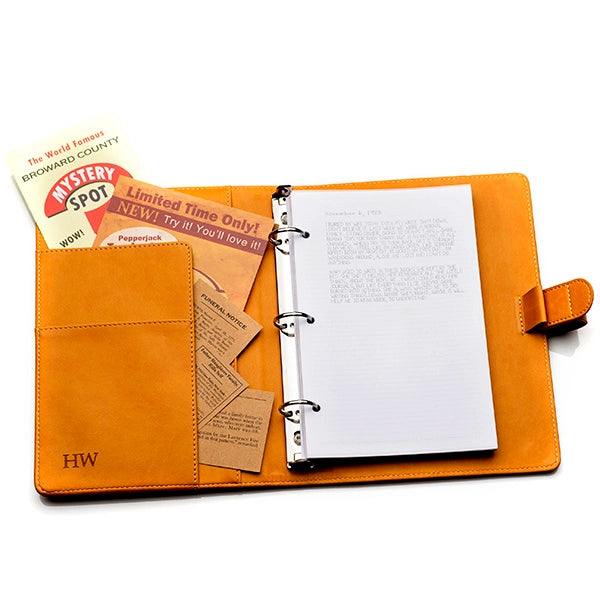 Top view of a brown faux leather journal, open against a white background. On the right side is a stack of white paper. On the left is a pocket filled with newspaper articles and advertisements.