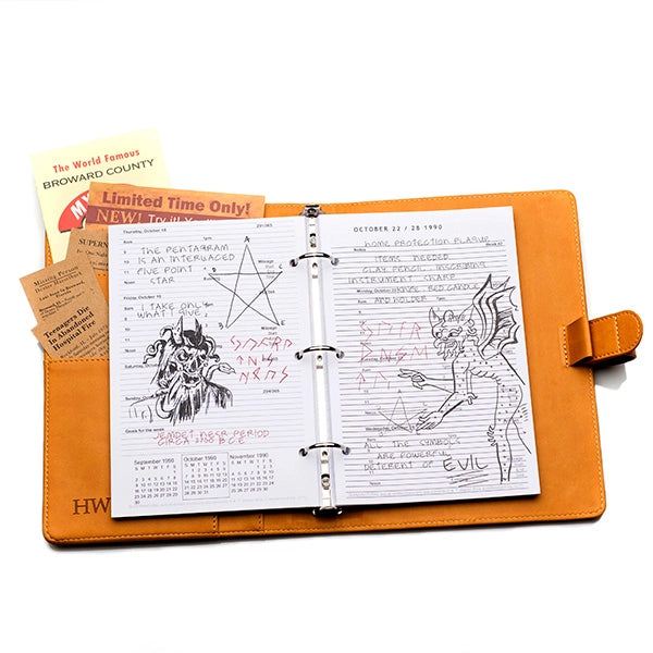 Top view of a brown faux leather journal, open against a white background. Spread through the center is a stack of white paper, with descriptions and drawings of demons and other monsters. Behind the paper on the left is a pocket filled with newspaper articles and advertisements.