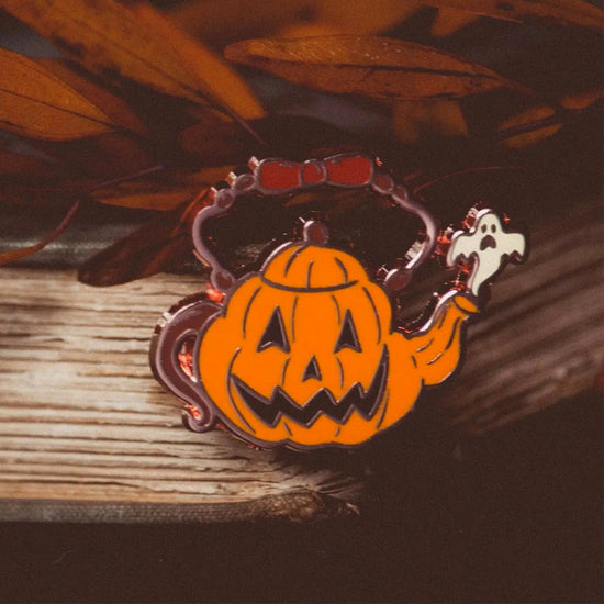 Close up view of an enamel pin depicting a teapot in the shape of an order jack-o-lantern, with a tiny white ghost coming out the spout. The pin is sitting on a wooden plank surrounded by orange leaves.
