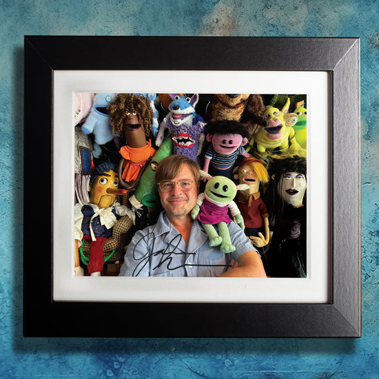 A black framed photograph against a blue wall. The photograph depicts puppeteer Jamie Shannon, surrounded by many of his puppet creations. The photograph is autographed by Jamie.