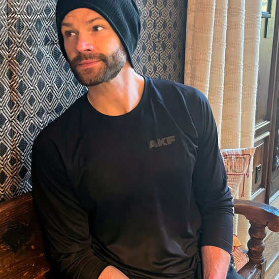 An image of actor Jared Padalecki sitting on a wooden bench, against a wall with a blue and white diamond pattern. Jared is wearing a black T-shirt with the letters AKF in dark grey on the left side.