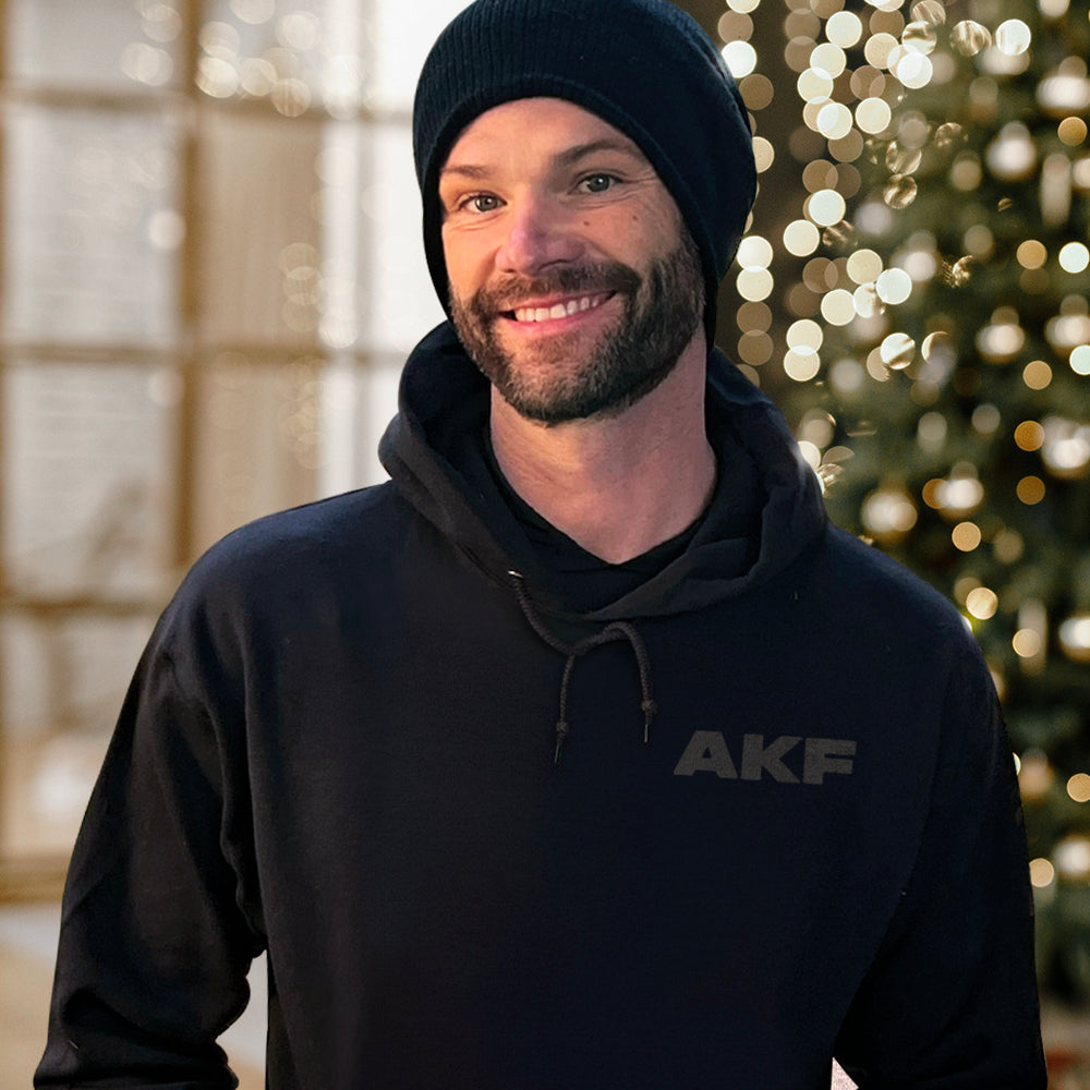 An image of actor Jared Padalecki standing in front of a Christmas tree with white lights. Jared is wearing a black pullover hoodie with the letters AKF in dark grey on the left side.