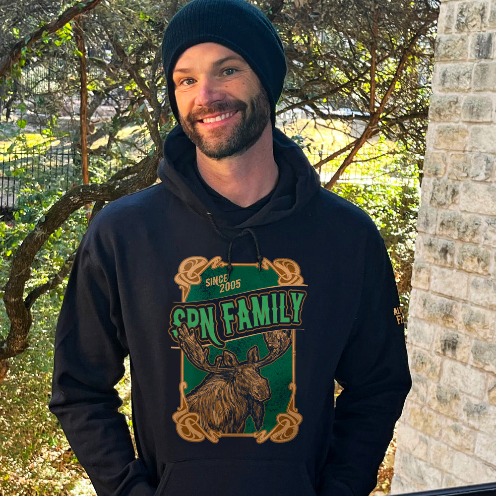 An image of actor Jared Padalecki wearing a black hooded sweatshirt. On the front of the hoodie is a green and brown rectangle with a brown frame. A brown moose is depicted at the bottom. Green text says "SPN Family" across the middle. At the top is light brown text saying "Since 2005." At the bottom right corner is a close up of the left sleeve, with yellow text saying "Always Keep Fighting." Behind Jared are trees and a brick wall.