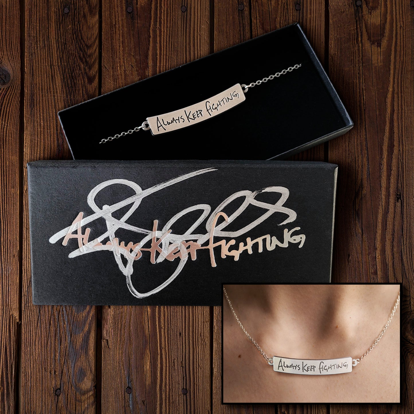 A silver bar necklace engraved with 'Always Keep Fighting' is displayed inside and on top of black jewelry boxes with metallic script lettering, set against a dark wood plank background. An inset image in the bottom right shows the necklace worn on a person's neck.
