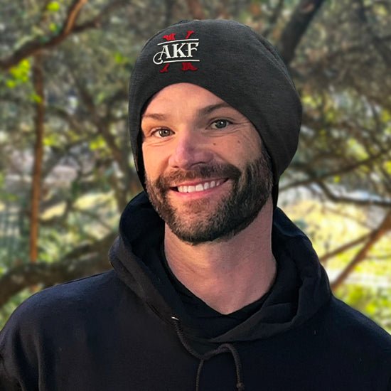 An image of actor Jared Padalecki wearing a black slouch beanie. The front of the beanie has a large red X, underneath the letters AKF in white. Behind Jared is a large tree.