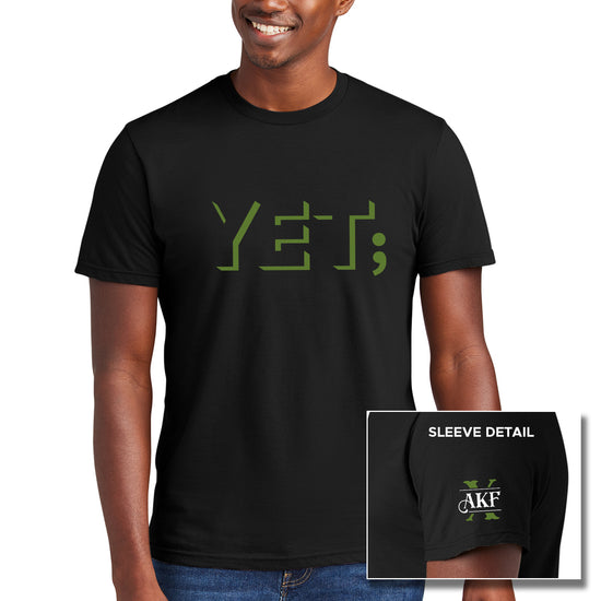 A male model wearing a black tee shirt. On the front of the shirt is the word "yet" in green block letters, with a semicolon at the end. An inset image of the left sleeve is at the bottom right corner. showing a large green X with the letters AKF above it on white.