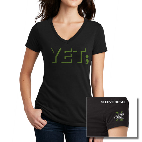 A female model wearing a black v-neck shirt. On the front of the shirt is the word "yet" in green block letters, with a semicolon at the end. An inset image of the left sleeve is at the bottom right corner. showing a large green X with the letters AKF above it on white.