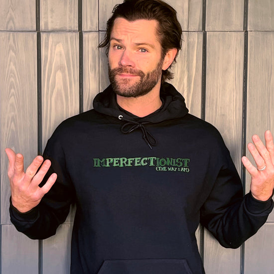 An image of actor Jared Padalecki wearing a black pullover hoodie. The front of the hoodie reads "IMPERFECTIONIST". The word "perfect" is printed in a darker shade, and the words "the way I am" are printed beneath it, so the full impression reads 'Imperfectionist / Perfect the way I am." Behind Jared is a wooden wall. 