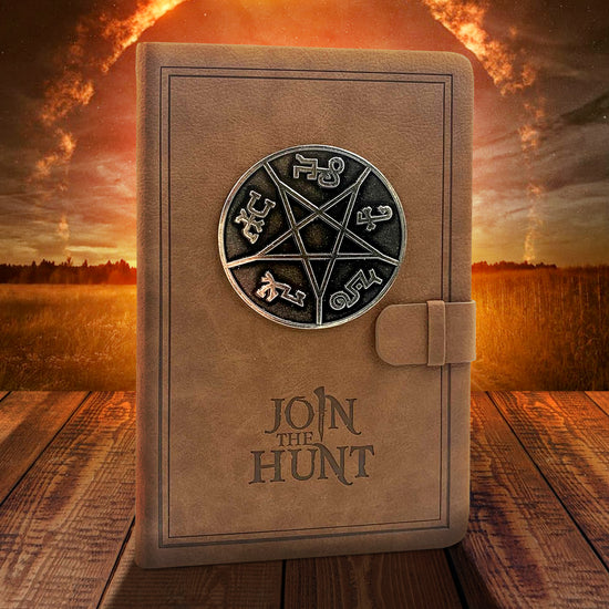 A brown, leathery notebook on a wooden table. On the cover is a Devil's Trap symbol in black and silver. At the bottom is dark brown text that says "Join the hunt." At the right side is a strap and loop to keep the notebook closed. In the background is a farm in bright sunlight, with a fiery ring in the sky.