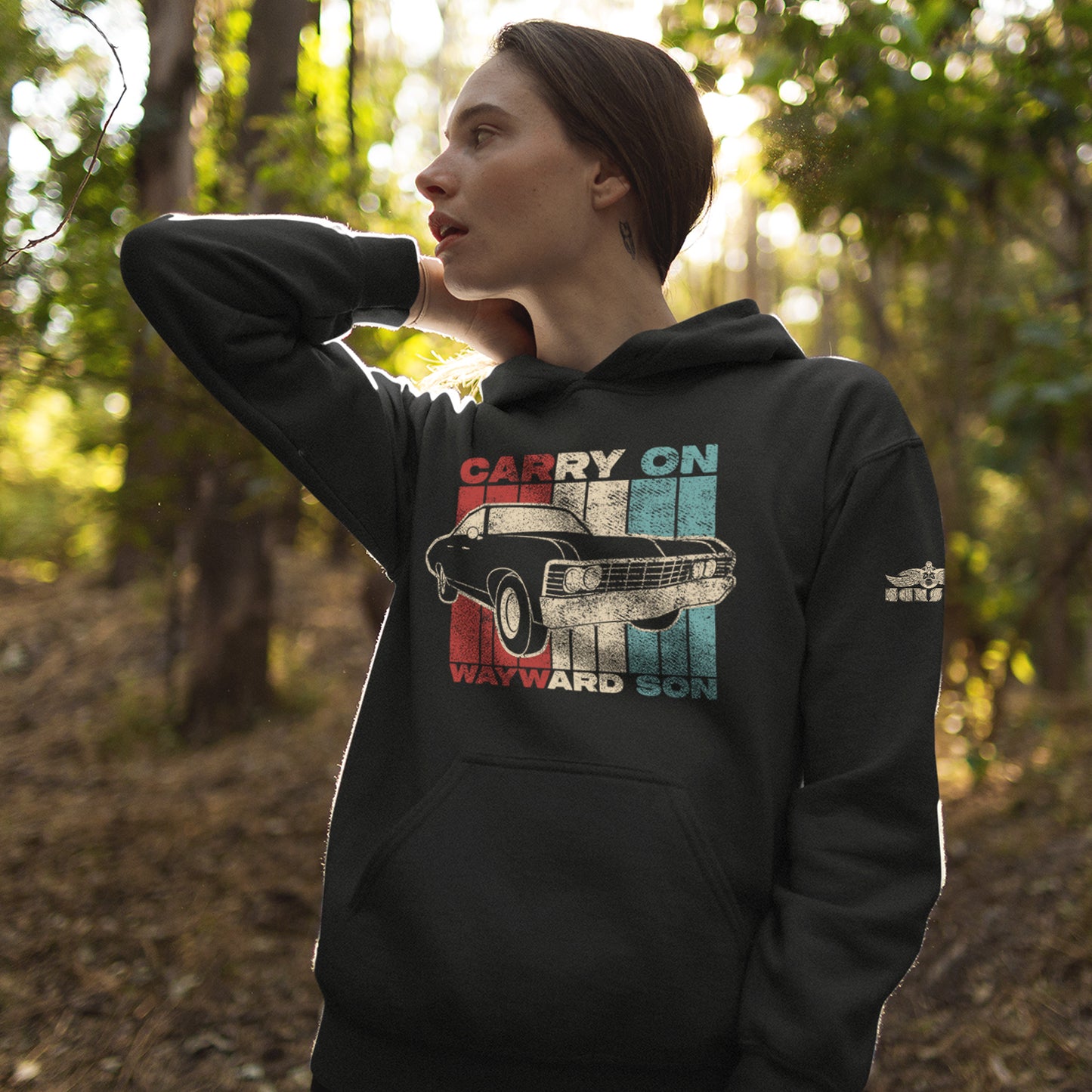 A female model standing in a forest and wearing a black hoodie. The hoodie features a flag comprised of red, white, and, blue vertical stripes. On the top and bottom of the flag are the words "CARRY ON WAYWARD SON" in the synced colored of the flag. A 1967 Impala is overlaid on top of the flag. On the left bicep is a small white Kansas Band logo.  
