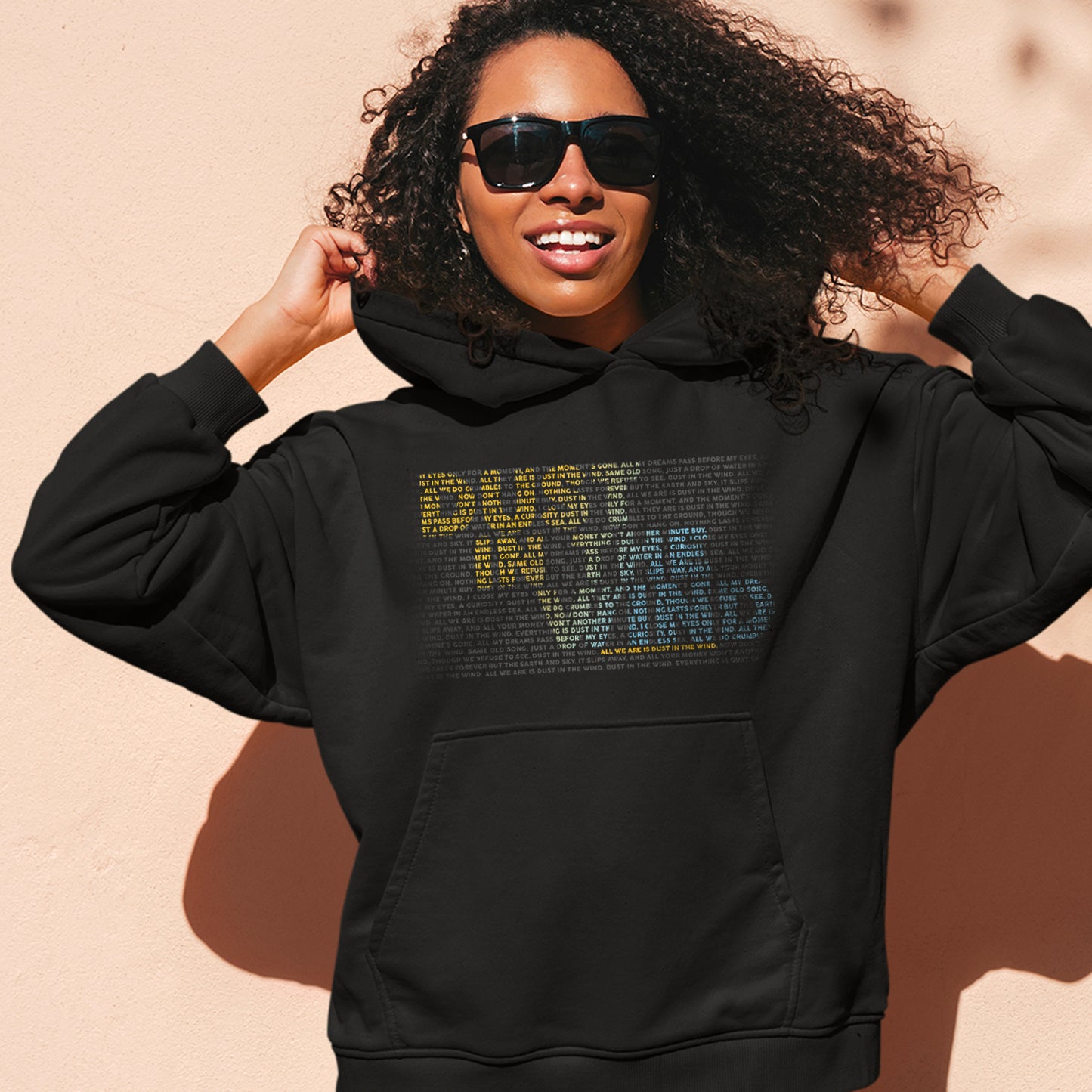 A female model wearing a black hoodie standing in front of a peach-colored wall. On the front of the hoodie is text saying "dust in the wind." The text is comprised of smaller text, colored yellow and blue, to form the dust in the wind phrase. 
