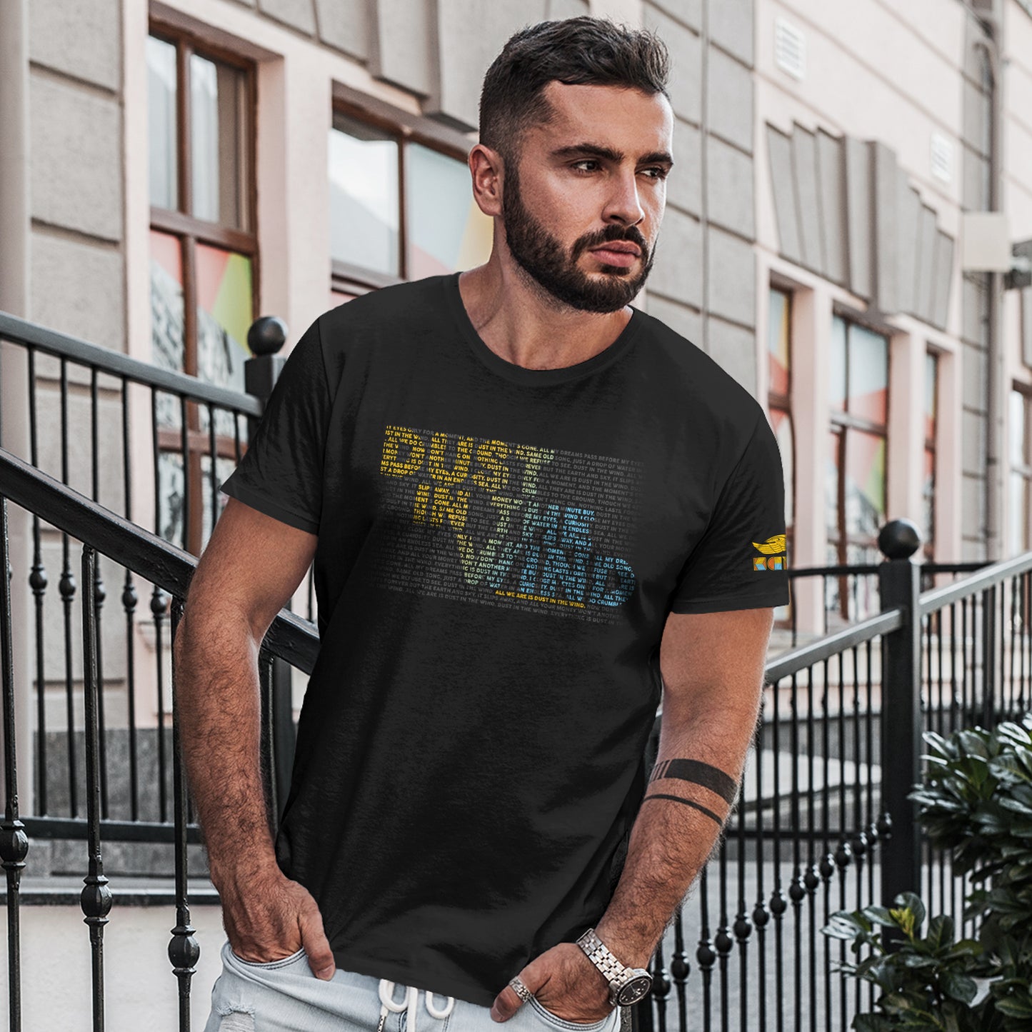 A male model wearing a black t-shirt standing in front of a grey building. On the front of the shirt is text saying "dust in the wind." The text is comprised of smaller text, colored yellow and blue, to form the dust in the wind phrase. On the left sleeve is the logo of the band Kansas.