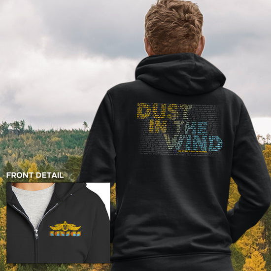 The back view of a male model wearing a black hoodie standing in a field. On the back side of the hoodie is text saying "dust in the wind." The text is comprised of smaller text, colored yellow and blue, to form the dust in the wind phrase. On the front of the hoodie is the logo for the band Kansas. 