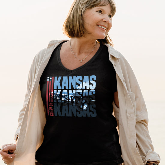 A female model wearing a black tee shirt, with an open white button down shirt over it. In the front of the shirt is blue text that says “Kansas Kansas Kansas,” with a black Chevy Impala superimposed on the text. On the left of the text are three red stripes with red text saying “carry on.”