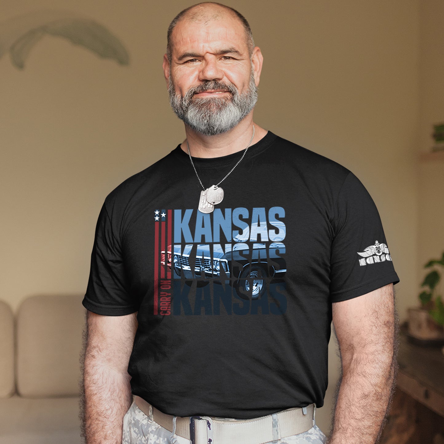 A male model wearing a black tee shirt. In the front of the shirt is blue text that says “Kansas Kansas Kansas,” with a black Chevy Impala superimposed on the text. On the left of the text are three red stripes with red text saying “carry on.”