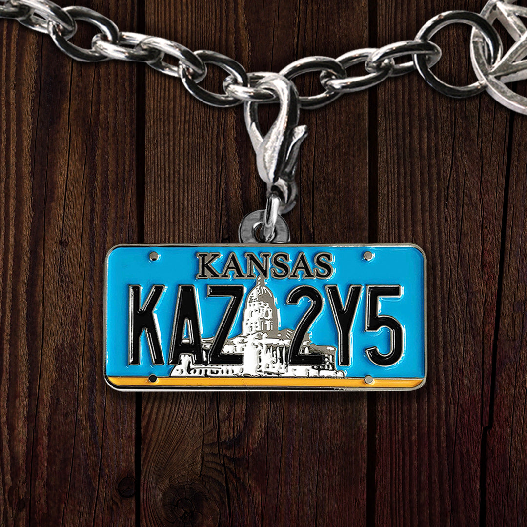 A bracelet charm hanging on a silver chain, against a dark wood background. The charm depicts alight blue  Kansas license plate,  with a white state building in the background. Black text says KANSAS: KAZ 2Y5