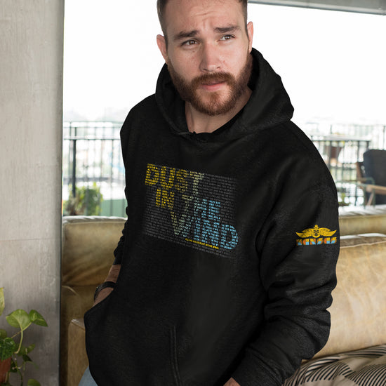 A male model wearing a black hoodie standing in a neutrally-themed apartment. On the front of the hoodie is text saying "dust in the wind." The text is comprised of smaller text, colored yellow and blue, to form the dust in the wind phrase.