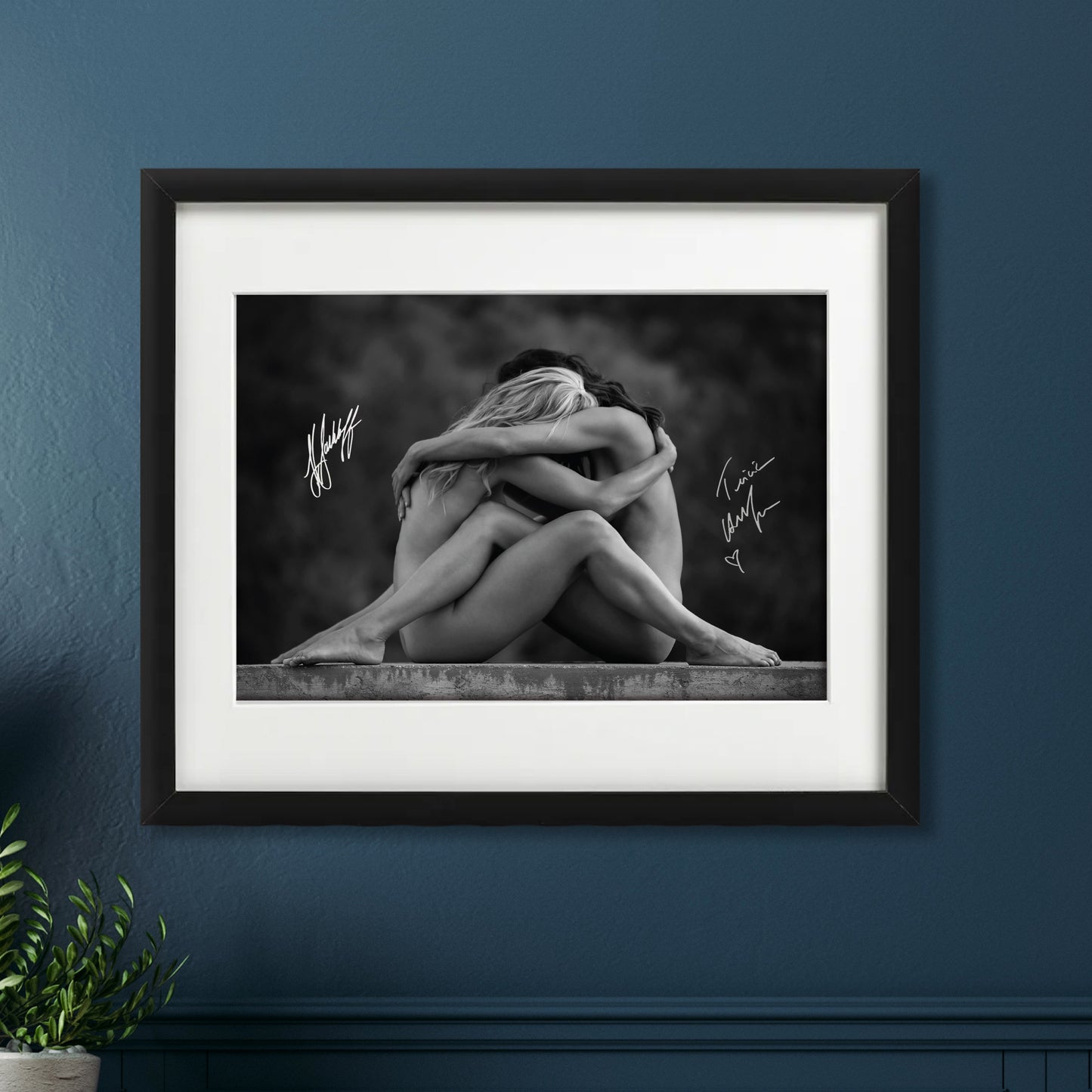 A black and white print with a black frame, against a dark blue wall. The print depicts the actors Katee Sackhoff and Tricia Helfer, sitting on a stone platform, facing each other. The actors are both naked, and are embracing each other, against a smoky black backdrop. On either side of them are the actor's autographs.