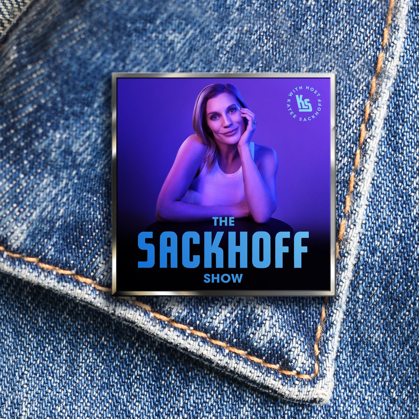 A dark blue enamel pin attached to a the collar of a denim jacket. The pin depicts actor Katee Sackhoff in a white tank top, holding her head in one hand. Blue text at the bottom says "The sackhoff show."