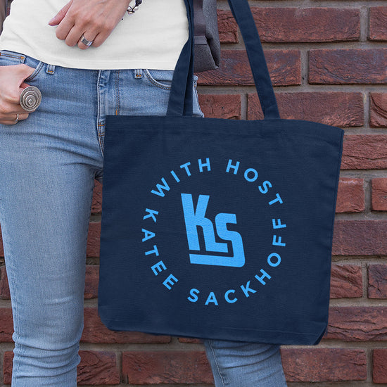 A model holding a dark blue cotton tote bag. On the front are the letters K and S in large light blue font. Surrounding the letters in a circle is light blue text saying "with host katee sackhoff."