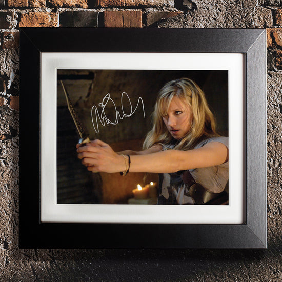 A black framed photograph against a brick and concrete wall. The photograph depicts actor Katie Cassidy, portraying the character Ruby from the series "Supernatural." Katie is wielding a dagger, with lit candles behind her. On the top left side is Katie's autograph in white ink.