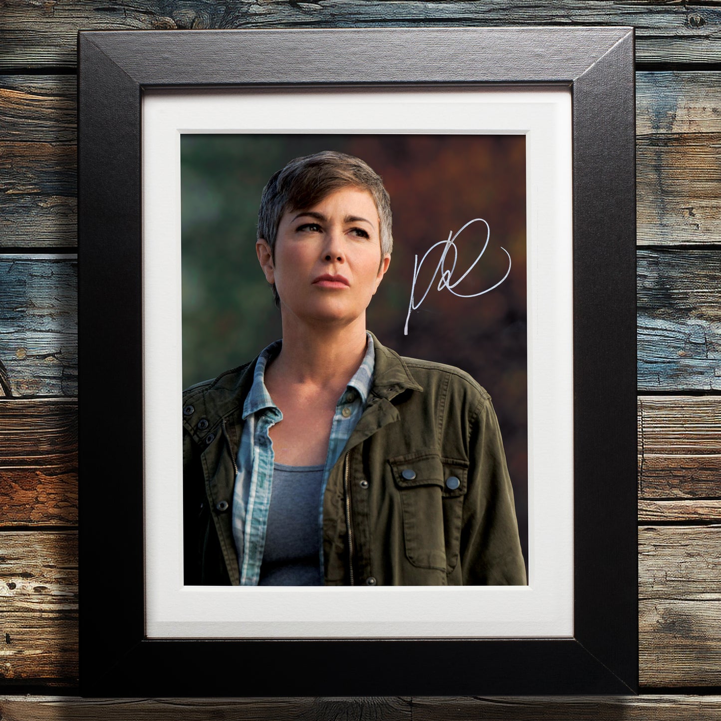 A black framed photograph against a wood. The photograph depicts actor Kim Rhodes, portraying the character Sheriff Jody Mills from the series "Supernatural." Kim is standing in front of indistinct trees with colorful leaves, dressed in a green jacket and blue shirt. In the top right corner is Kim's autograph in white ink.