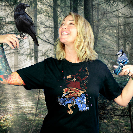 An image of the actress Kim Rhodes standing in front of a desolate forrest. She's wearing a black T-shirt with a drawing of two crows on the front, one in red, the other in blue. At the center in red text is "murder party." Two birds are sitting on each of Kim's outstretched pointer fingers.
