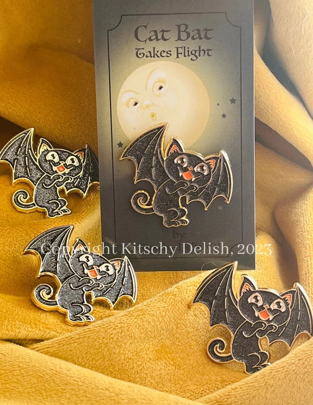 Three enamel lapel pins of a cute black cat bat clinging to a dark card labeled 'Cat Bat Takes Flight,' arranged on draped mustard-yellow fabric.