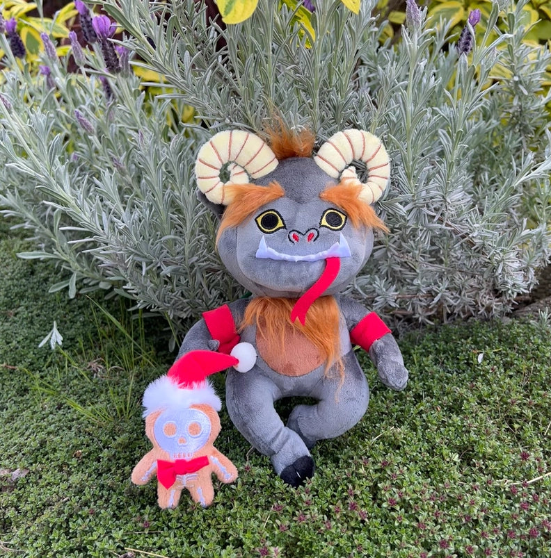 A grey plushie depicting the mythological figure Krampus. At the top of his head are yellow horns and patches of brown hair. Across his face are white fangs, and a long red tongue. A small gingerbread figure wearing a santa hat is on the plushie's back.Behind the plushies is a flowery bush.